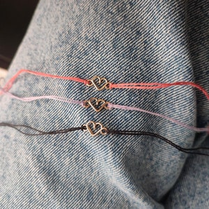 Könnte beinhalten: Vier Armbänder mit einem goldenen Herz-Anhänger. Die Armbänder sind in verschiedenen Farben: rosa, lila, schwarz und weiß.