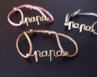 May include: Three bracelets with the word "papa" spelled out in gold wire. The bracelets are on a black surface. The bracelets are pink, brown, and green.