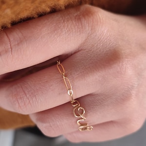 Könnte beinhalten: Ein goldener Kettenring mit einem Ring mit einem kursiv geschriebenen Namen am selben Finger. Der Namensring sagt "Love".