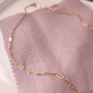 May include: A gold chain necklace with a delicate, oval-shaped link design. The chain is lying on a soft, pink fabric surface.
