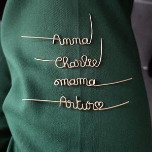May include: Four gold wire names, Anna, Charlie, mama, and Arthur with a heart, are displayed on a green fabric background.