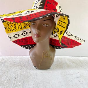 May include: A wide-brimmed sun hat with a colorful geometric pattern in red, yellow, black, and white. The hat is made of fabric and has a wide brim that provides shade from the sun.