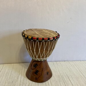 May include: A small, brown wooden djembe drum with a natural leather head. The drum has a carved design on the base and is decorated with a red, yellow, and black rope around the top.