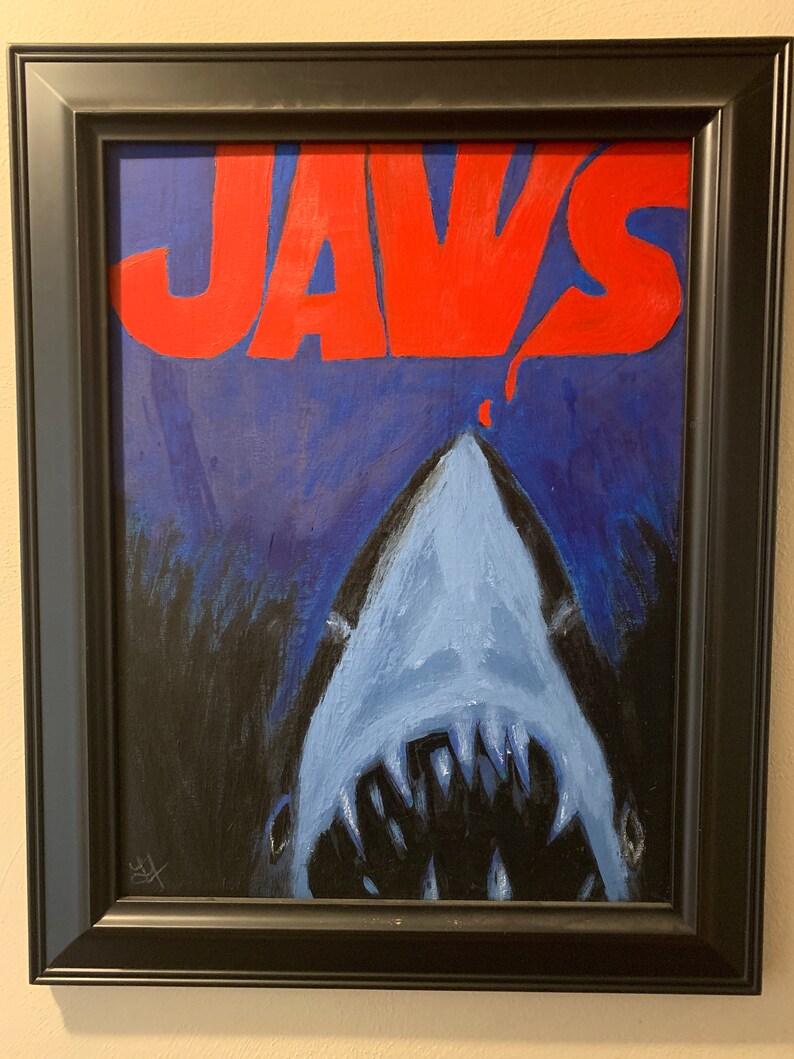Op de afbeelding: Ingelijst schilderij met het woord "JAWS" in rode letters boven de geopende bek van een blauwe haai. De haai is geschilderd in blauw- en wittinten tegen een donkerblauwe achtergrond. Het kunstwerk is ingelijst in een zwarte lijst.