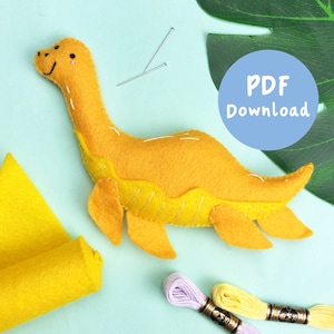 May include: A yellow felt dinosaur toy with a friendly face, embroidered details, and a PDF download button. The toy is surrounded by craft supplies, including felt squares, needles, and embroidery floss.
