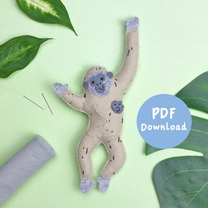 Può includere: Un giocattolo di scimmia di peluche beige in feltro con un cucciolo di scimmia sul petto. La scimmia ha un viso sorridente con occhi neri e un naso rosa. Il giocattolo è su uno sfondo verde con foglie verdi e un cerchio blu con il testo "Download PDF".