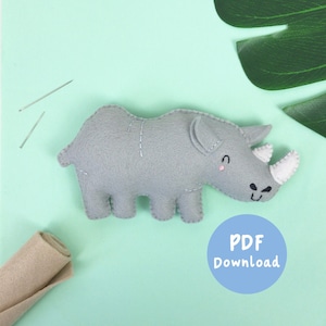 May include: A gray felt rhinoceros stuffed animal with white felt horn and stitched details. The rhino is lying on a light green surface with two sewing pins and a roll of beige felt. A blue circle with the text "PDF Download" is in the lower right corner.