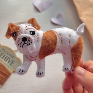 May include: A small, handcrafted felt bulldog ornament featuring white and tan patches, stitched facial details, and a short tail. The figure is made from soft felt material with visible embroidery thread accents defining the dog's features.