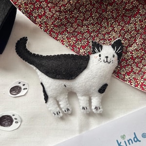 Könnte beinhalten: Ein handgefertigtes Filz-Katzenornament mit weißem Körper und schwarzen Flecken auf Rücken, Schwanz und Kopf. Die Katze hat gestickte Gesichtszüge und zwei separate kleine weiße Filz-Pfotenabdrücke mit schwarzen Ballen auf der linken Seite.
