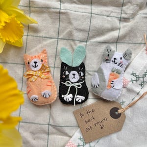 May include: Three handmade felt cat ornaments in orange, black, and grey. The orange cat has a yellow bow, the black cat has mint green bunny ears, and the grey cat holds a carrot. A tag reads "to the best cat mum."