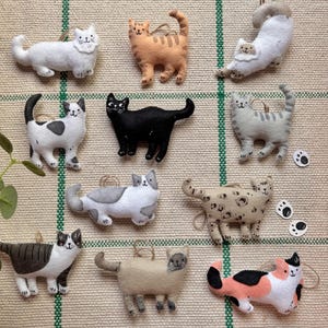 May include: A collection of handmade felt cat ornaments in various colors and patterns. The cats include white, orange, black, gray, and calico designs. Each ornament has a loop for hanging and is displayed on a textured, neutral background.