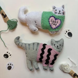 Könnte beinhalten: Zwei handgefertigte Katzen-Spielzeuge aus Filz. Eine weiße Katze trägt einen grünen Pullover mit einem Herz mit der Aufschrift "Cool Cat". Die andere ist grau mit schwarzen Streifen und einem rosa Pullover mit schwarzen Wellenlinien. Pfotenabdruck-Ausschnitte und Garnrollen sind verstreut.