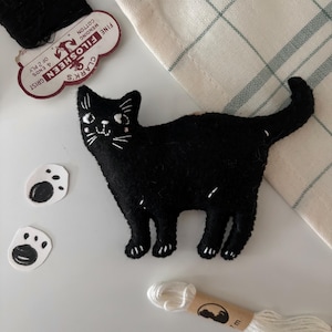 Peut inclure: Un chat noir en feutre avec des détails brodés en blanc, notamment des moustaches et des traits du visage. Deux découpes de pattes et une bobine de fil blanc sont également visibles. Le chat est le point central, sur un fond neutre.