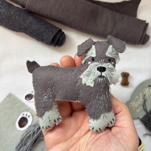 Può includere: Figurina di cane Schnauzer fatta a mano in feltro nei toni del grigio e dell'azzurro. Il cane ha dettagli ricamati, tra cui naso e baffi neri. Un piccolo pezzo di feltro a forma di osso marrone è accanto al cane.
