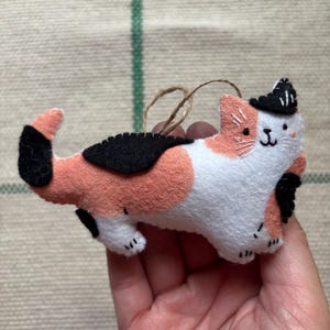 May include: A handmade felt cat ornament in white, peach, and black. The cat has a small black hat and a string for hanging. The cat's body is white with peach and black spots. The ornament is held in a hand.