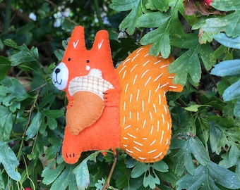 May include: A felt orange squirrel holding a brown acorn. The squirrel has white stitching for detail. The squirrel is sitting on a green leafy bush.