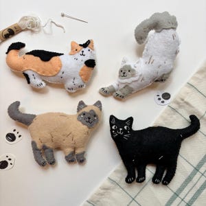 May include: Four handmade felt cat figures in various poses and colors. One is calico, another is gray and white, a third is tan and gray, and the last is solid black. The cats are on a white surface with sewing supplies and paw print stickers.