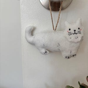 May include: A white felt cat ornament with black paw details, whiskers, and a smiling face. The cat is suspended by a natural twine string. The ornament is hanging from a silver doorknob.