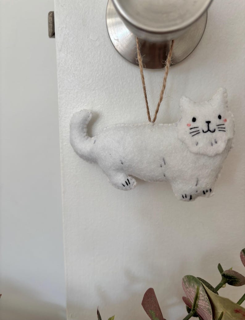 May include: A white felt cat ornament with black paw details, whiskers, and a smiling face. The cat is suspended by a natural twine string. The ornament is hanging from a silver doorknob.