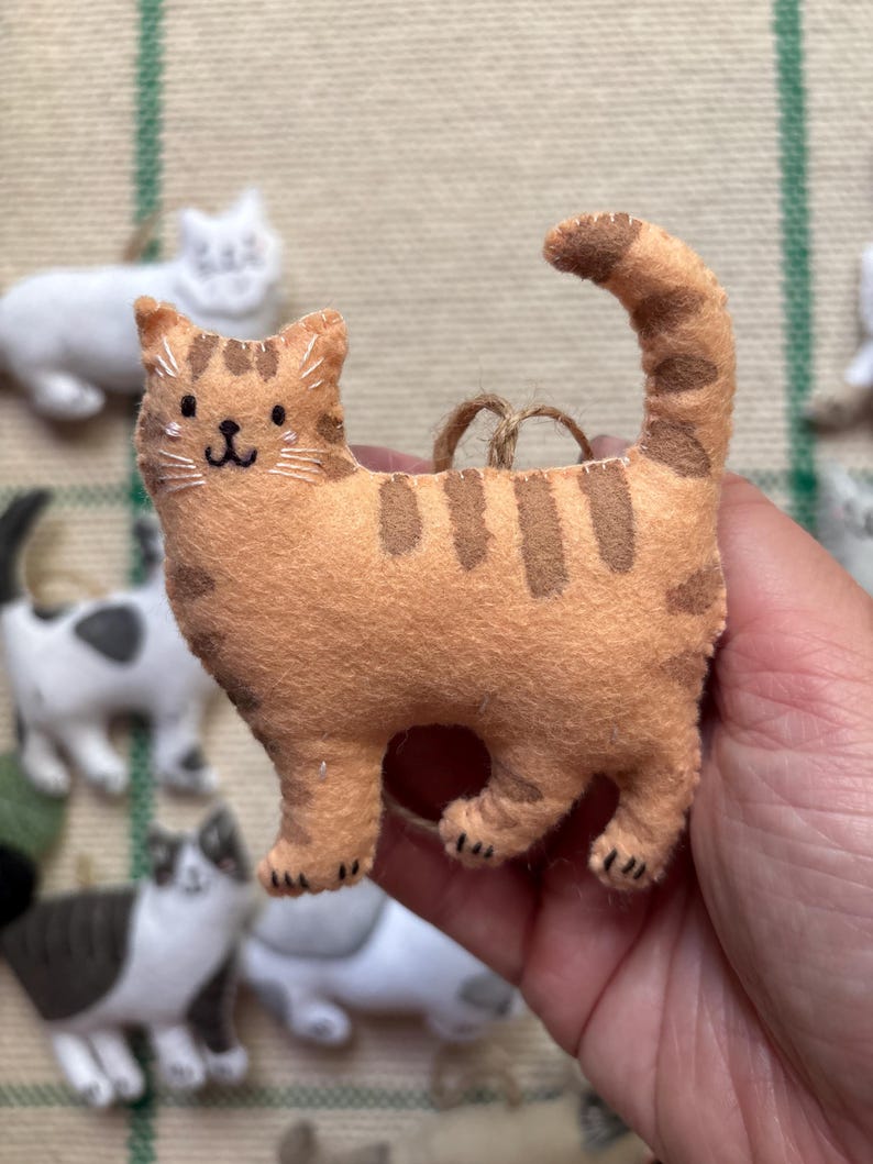 May include: A handmade orange tabby cat ornament with brown stripes and a smiling face. The felt cat has a curved tail and a jute string for hanging. Other cat ornaments are visible in the background.