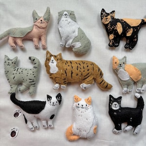 May include: A selection of handmade felt cat figurines in diverse colours and patterns. The cats feature calico, tabby, and solid-coloured designs. Each cat has embroidered details, including facial features and markings. The figurines are arranged on a white surface.