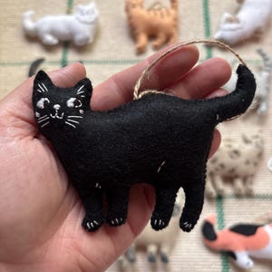 May include: A black felt cat ornament with white embroidered details, including eyes, whiskers, and paws. The cat has a looped twine hanger. Other cat ornaments in various colors are in the background.