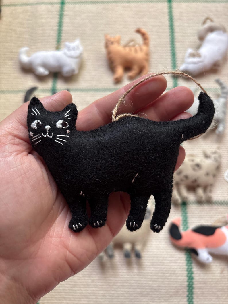 May include: A black felt cat ornament with white embroidered details, including eyes, whiskers, and paws. The cat has a looped twine hanger. Other cat ornaments in various colors are in the background.