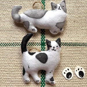 May include: Two handmade felt cat ornaments. One is white with gray spots and the other is white with black spots. Both have a loop for hanging. Two small paw print cutouts are also included.