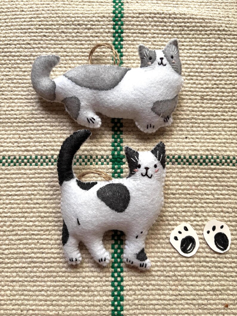 May include: Two handmade felt cat ornaments. One is white with gray spots and the other is white with black spots. Both have a loop for hanging. Two small paw print cutouts are also included.