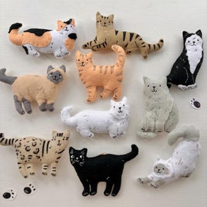Puede incluir: Una colección de figuras de gatos de fieltro hechas a mano en varios colores y patrones. Los gatos incluyen calicó, atigrados, siameses y gatos de un solo color. Cada gato tiene rasgos faciales bordados y está posado en diferentes posiciones.