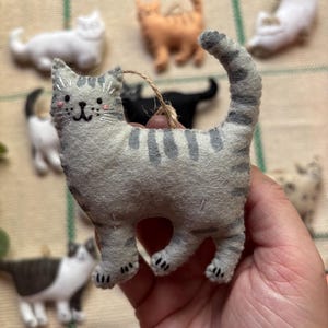 May include: A gray felt cat ornament with dark gray stripes and a smiling face. The cat has a jute string for hanging and is surrounded by other felt cat ornaments in various colors and poses.