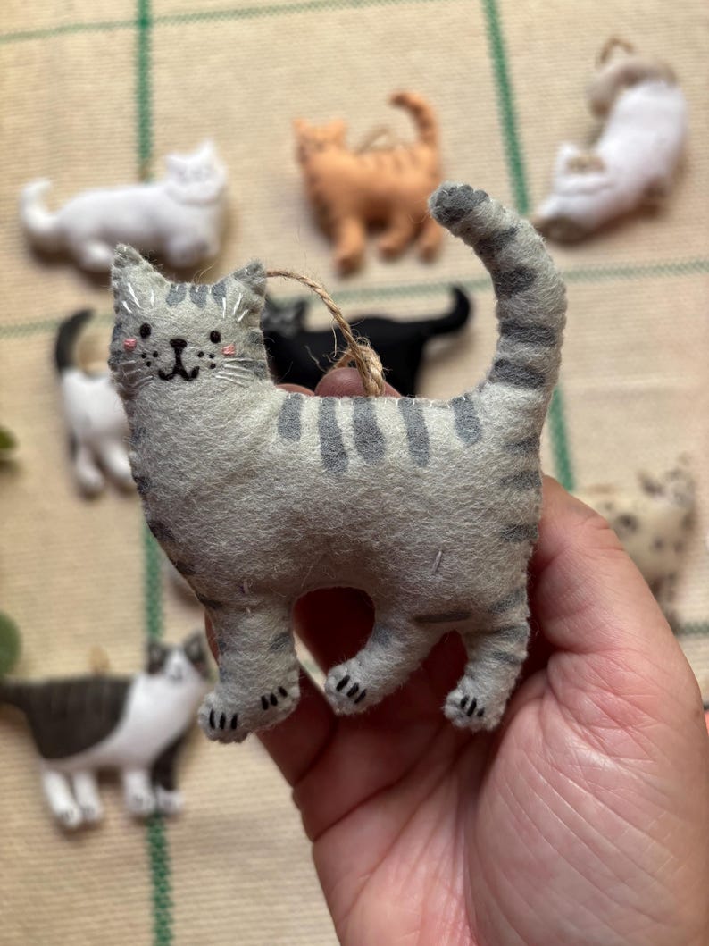 May include: A gray felt cat ornament with dark gray stripes and a smiling face. The cat has a jute string for hanging and is surrounded by other felt cat ornaments in various colors and poses.