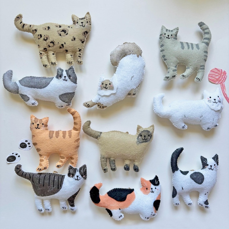 May include: A collection of handmade felt cat figurines in various colors and patterns. The cats include a spotted beige cat, a gray tabby, an orange tabby, and a white cat with gray spots. Some cats are posed in playful positions.