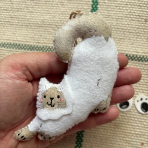 May include: A handmade felt cat ornament in a stretching pose. The cat is primarily white with tan accents on the head, paws, and tail. It has a smiling face with pink cheeks and black whiskers. A small loop of twine is attached for hanging.