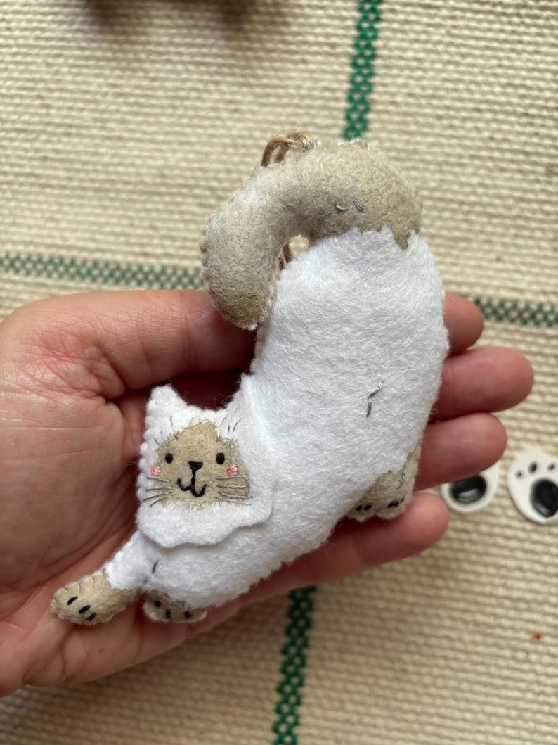 May include: A handmade felt cat ornament in a stretching pose. The cat is primarily white with tan accents on the head, paws, and tail. It has a smiling face with pink cheeks and black whiskers. A small loop of twine is attached for hanging.