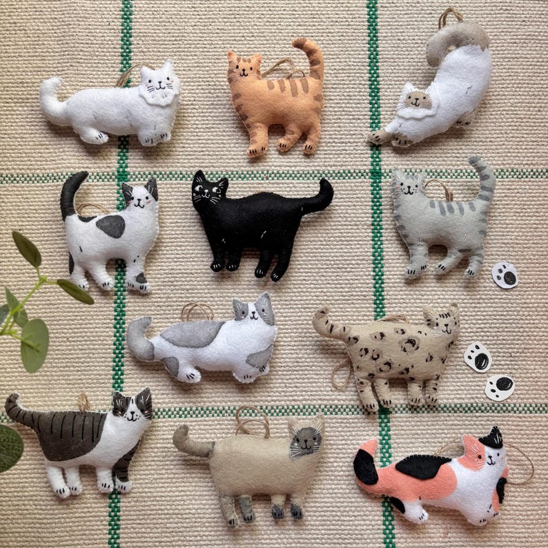 May include: A collection of handmade felt cat ornaments in various colors and patterns. The cats include white, orange, black, gray, and calico designs. Each ornament has a loop for hanging and is displayed on a textured, neutral background.