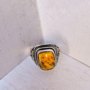 May include: A silver ring featuring a rectangular, mustard-yellow stone with dark speckles. The metal setting is decorated with a twisted rope border and intricate beaded detailing along the sides, framing the central gemstone securely.