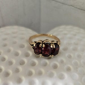 May include: A gold ring featuring three oval-cut deep red garnet gemstones arranged horizontally in a row. The stones are held in place by a gold prong setting, and the band is a simple, smooth gold metal design that encircles the finger.