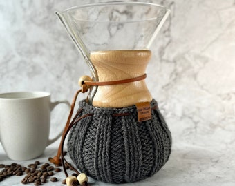 May include: A glass pour-over coffee maker with a light wood collar and a dark gray ribbed knit cozy. The cozy features a brown leather tag labeled Red Cellar Designs and a leather cord with two light wood beads. A white mug and coffee beans.