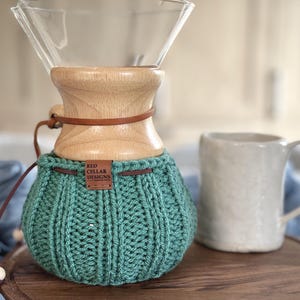 K&ouml;nnte beinhalten: Ein Glas-Kaffeebereiter mit einem hellen Holzkragen und einem braunen Lederband. Der untere Teil ist mit einem gerippten, salbeigr&uuml;nen H&auml;kel&uuml;berzug bedeckt, der ein kleines braunes Lederetikett mit der Aufschrift RED CELLAR DESIGNS tr&auml;gt.
