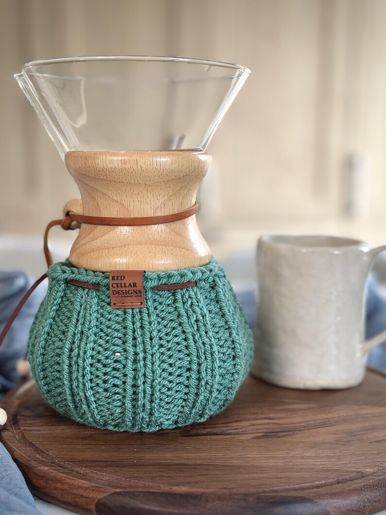 K&ouml;nnte beinhalten: Ein Glas-Kaffeebereiter mit einem hellen Holzkragen und einem braunen Lederband. Der untere Teil ist mit einem gerippten, salbeigr&uuml;nen H&auml;kel&uuml;berzug bedeckt, der ein kleines braunes Lederetikett mit der Aufschrift RED CELLAR DESIGNS tr&auml;gt.