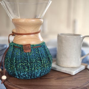 K&ouml;nnte beinhalten: Ein Glas-Kaffeebereiter mit einem hellen Holzkragen und einem dunkelbraunen Lederband. Der untere Teil ist mit einem gerippten, handgestrickten &Uuml;berzug in verschiedenen Blau-, Gr&uuml;n- und T&uuml;rkist&ouml;nen bedeckt, der ein braunes Etikett RED CELLAR DESIGNS tr&auml;gt.