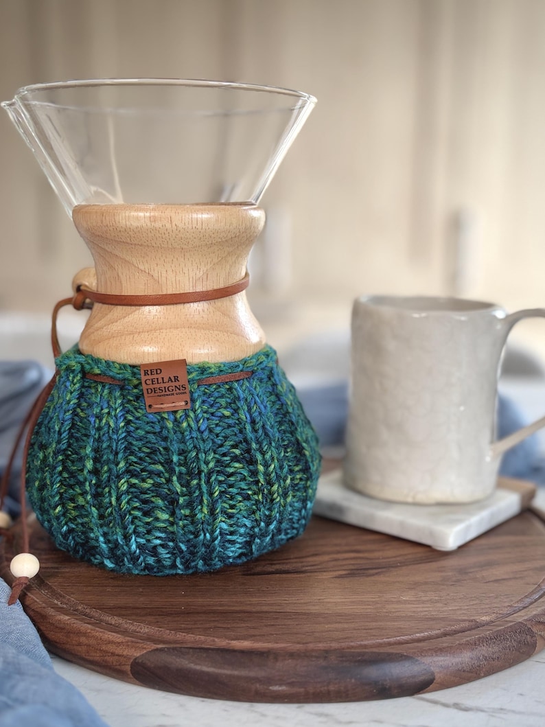 K&ouml;nnte beinhalten: Ein Glas-Kaffeebereiter mit einem hellen Holzkragen und einem dunkelbraunen Lederband. Der untere Teil ist mit einem gerippten, handgestrickten &Uuml;berzug in verschiedenen Blau-, Gr&uuml;n- und T&uuml;rkist&ouml;nen bedeckt, der ein braunes Etikett RED CELLAR DESIGNS tr&auml;gt.