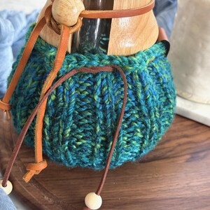 K&ouml;nnte beinhalten: Eine Glaskaffeekaraffe mit Holzkragen und einem handgestrickten, blau-t&uuml;rkisfarbenen Garn&uuml;berzug. Der &Uuml;berzug hat eine gerippte Textur und wird mit einem d&uuml;nnen braunen Lederband am Hals befestigt, das an den Enden kleine Holzperlen aufweist.