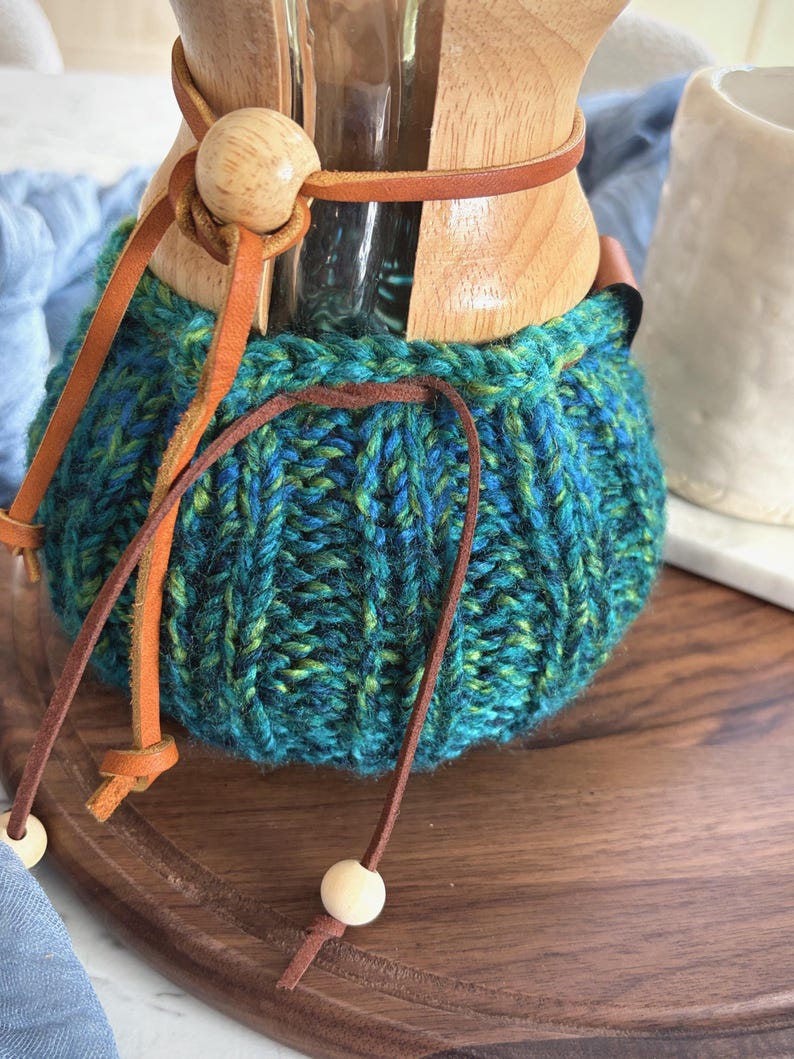 K&ouml;nnte beinhalten: Eine Glaskaffeekaraffe mit Holzkragen und einem handgestrickten, blau-t&uuml;rkisfarbenen Garn&uuml;berzug. Der &Uuml;berzug hat eine gerippte Textur und wird mit einem d&uuml;nnen braunen Lederband am Hals befestigt, das an den Enden kleine Holzperlen aufweist.