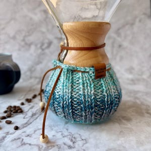 K&ouml;nnte beinhalten: Ein Kaffeebereiter aus klarem Glas mit einem hellen Holzkragen und einem blau-wei&szlig;en Strick&uuml;berzug im Ombr&eacute;-Stil. Ein braunes Lederband mit Holzperlen umschlie&szlig;t den Hals, und ein kleines braunes Etikett tr&auml;gt die Aufschrift Red Cellar Design.