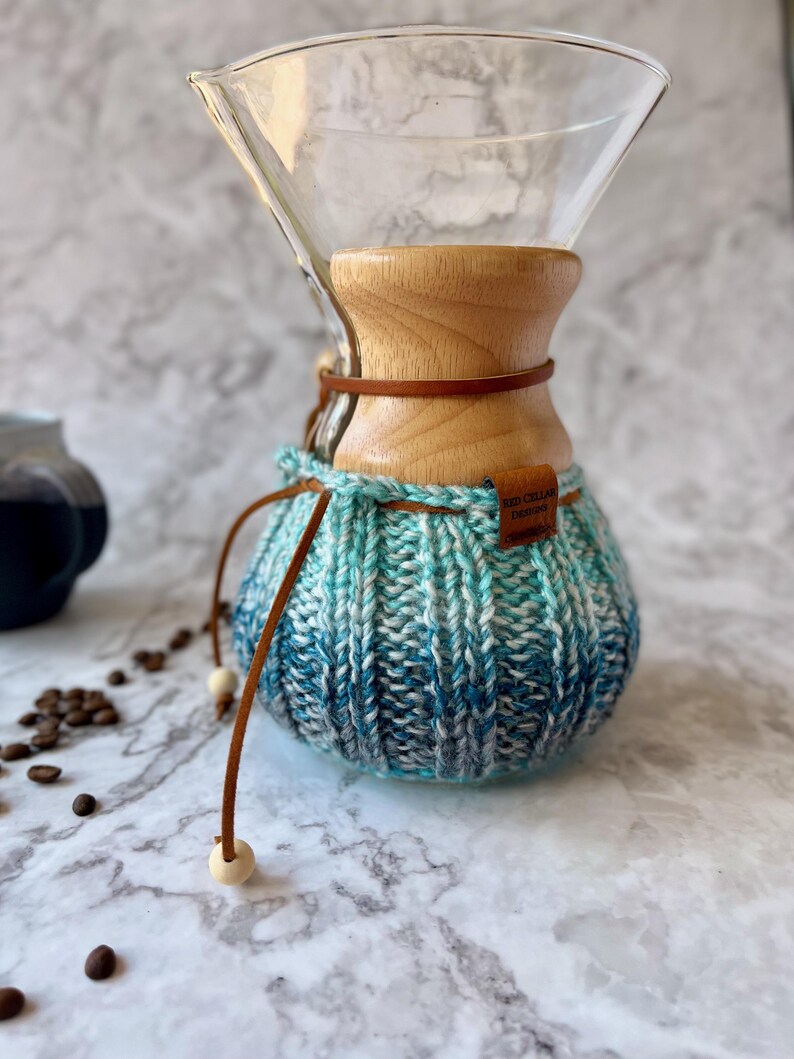 K&ouml;nnte beinhalten: Ein Kaffeebereiter aus klarem Glas mit einem hellen Holzkragen und einem blau-wei&szlig;en Strick&uuml;berzug im Ombr&eacute;-Stil. Ein braunes Lederband mit Holzperlen umschlie&szlig;t den Hals, und ein kleines braunes Etikett tr&auml;gt die Aufschrift Red Cellar Design.