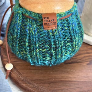 K&ouml;nnte beinhalten: Ein handgestrickter &Uuml;berzug in verschiedenen Blau- und Gr&uuml;nt&ouml;nen mit gerippter Textur und einer braunen Lederschnur mit Holzperlen. Ein rechteckiges braunes Lederetikett ist angebracht und zeigt den Text RED CELLAR DESIGNS HANDMADE GOODS.
