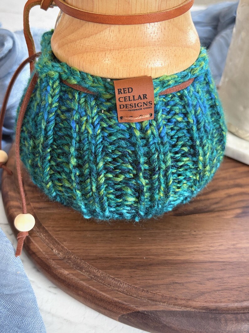 K&ouml;nnte beinhalten: Ein handgestrickter &Uuml;berzug in verschiedenen Blau- und Gr&uuml;nt&ouml;nen mit gerippter Textur und einer braunen Lederschnur mit Holzperlen. Ein rechteckiges braunes Lederetikett ist angebracht und zeigt den Text RED CELLAR DESIGNS HANDMADE GOODS.