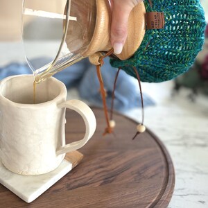 K&ouml;nnte beinhalten: Ein klarer Glas-Kaffeebereiter mit Holzkragen und einem blaugr&uuml;nen Strick&uuml;berzug, der ein braunes Etikett mit der Aufschrift RED CELLAR DESIGNS tr&auml;gt. Der Kaffeebereiter gie&szlig;t Fl&uuml;ssigkeit in eine wei&szlig;e Keramiktasse mit strukturierter Oberfl&auml;che.